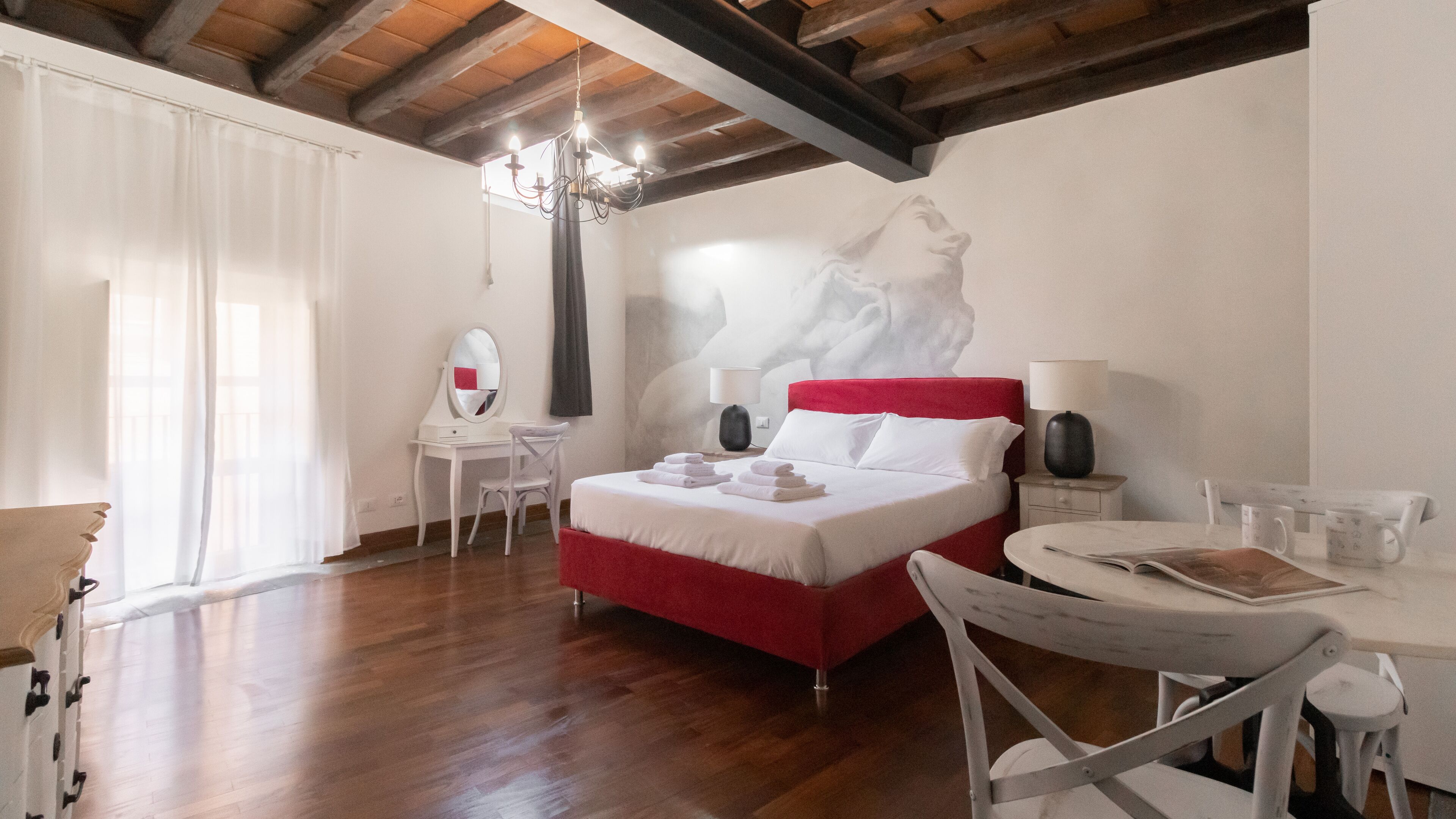 Camera d'albergo con soffitto in legno, un letto con testiera rossa, una scrivania bianca con uno specchio, un tavolino rotondo con una sedia e una lampada a parete.