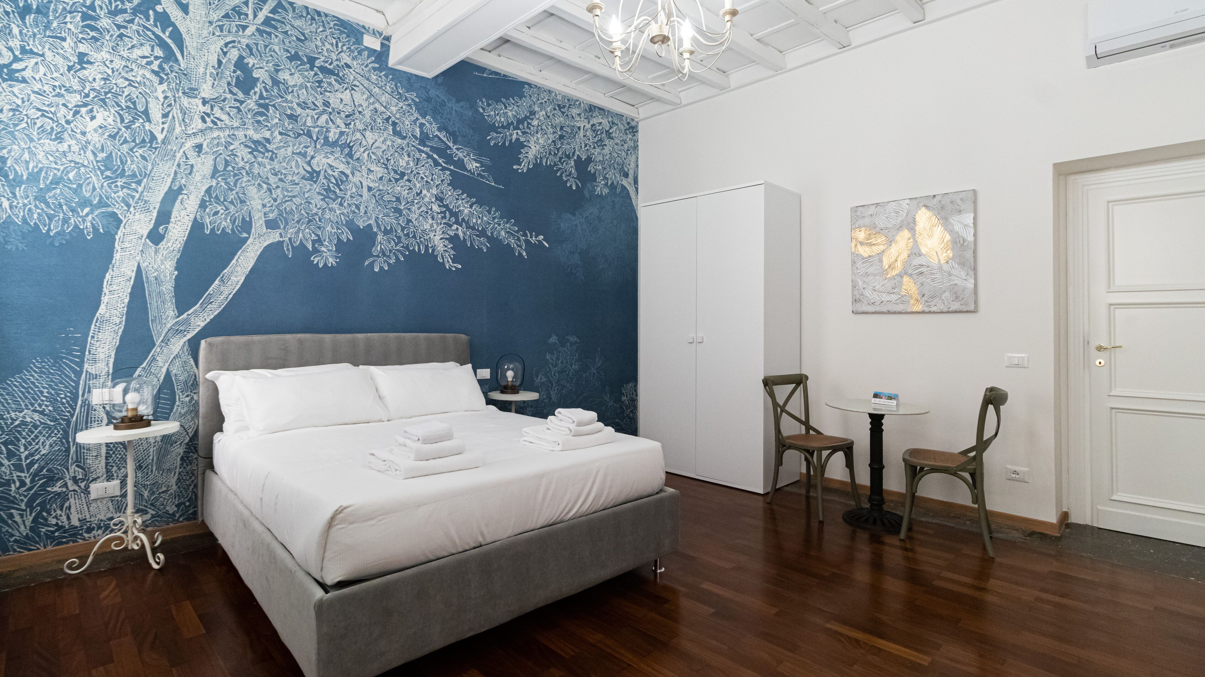 Ein Hotelzimmer mit einem großen Bett, einem kleinen Tisch mit Stühlen, einer Chandelierleuchtre und einer blauen Wand mit einem Baum-Wandgemälde.