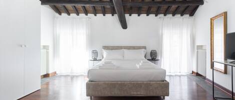 Un dormitorio amplio con una cama grande, vigas de madera en el techo, cortinas blancas y un televisor sobre un soporte.