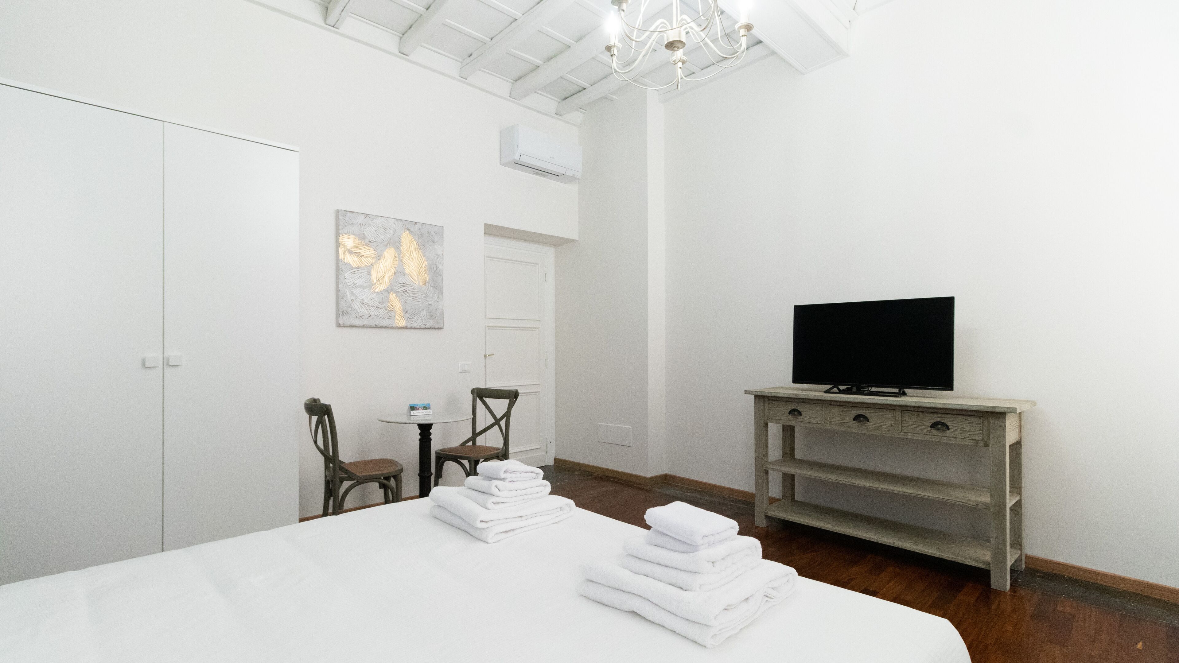 Een hotelkamer met een groot bed, een televisie op een houten standaard, een tafeltje met stoelen, en een schilderij aan de muur.