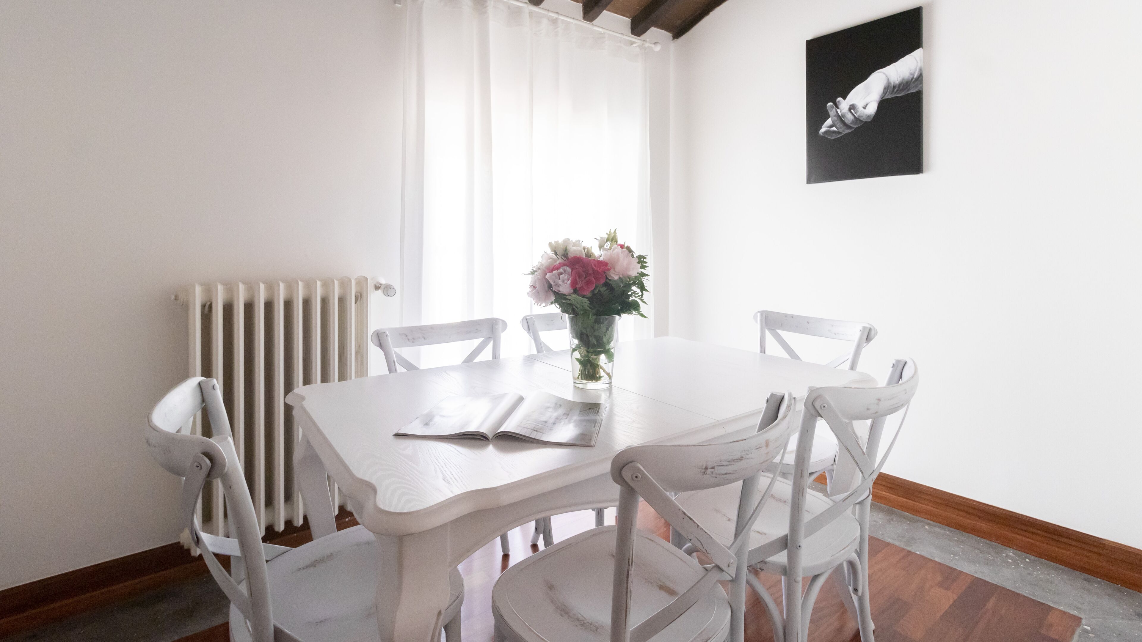 Una mesa de comedor blanca con cuatro sillas, un florero con flores, un radiador y un cuadro en la pared.