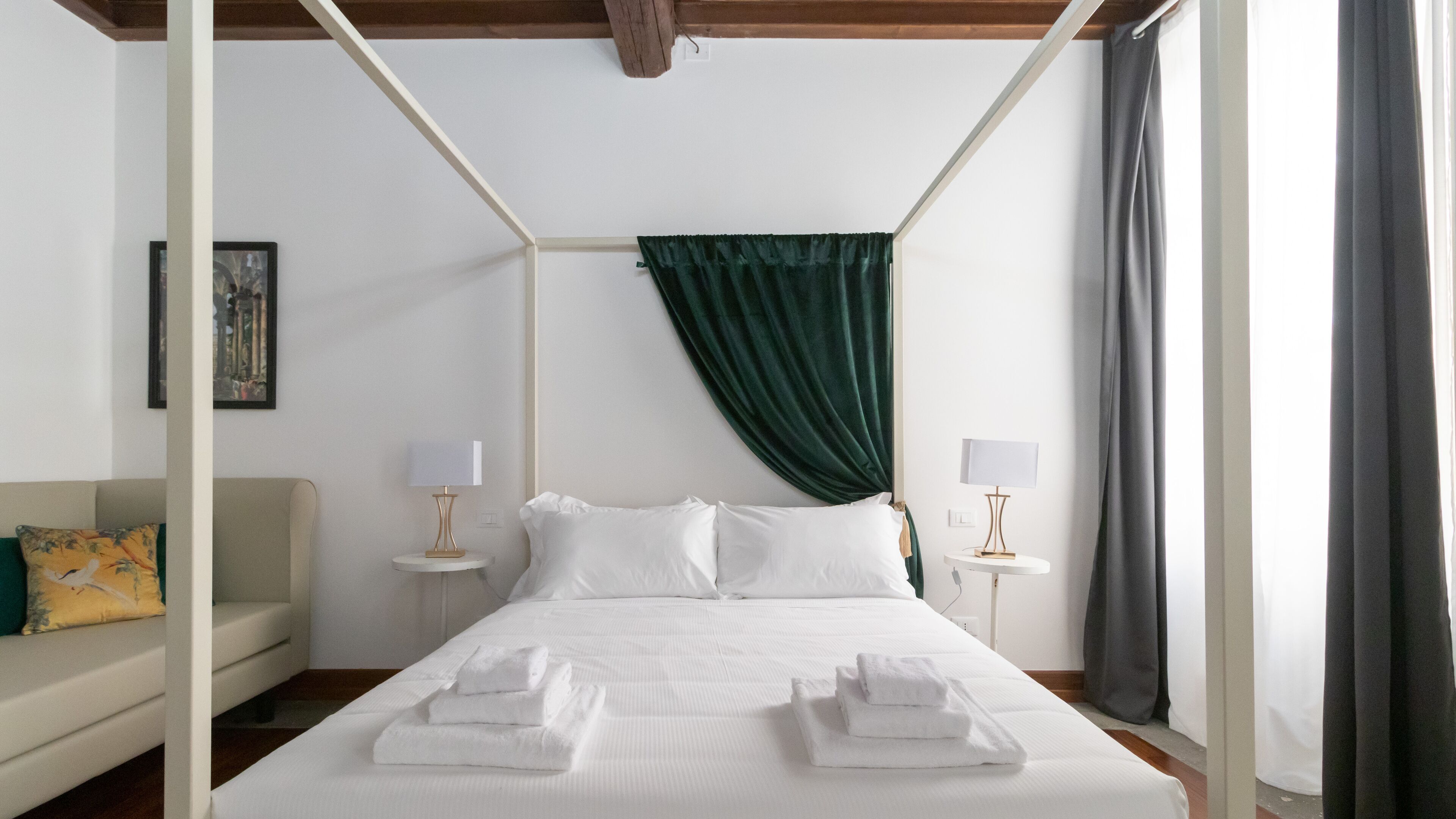 A four-poster bed with white linens and towels, two bedside tables with lamps, a green curtain, a framed picture on the wall, and a sofa with a yellow pillow.