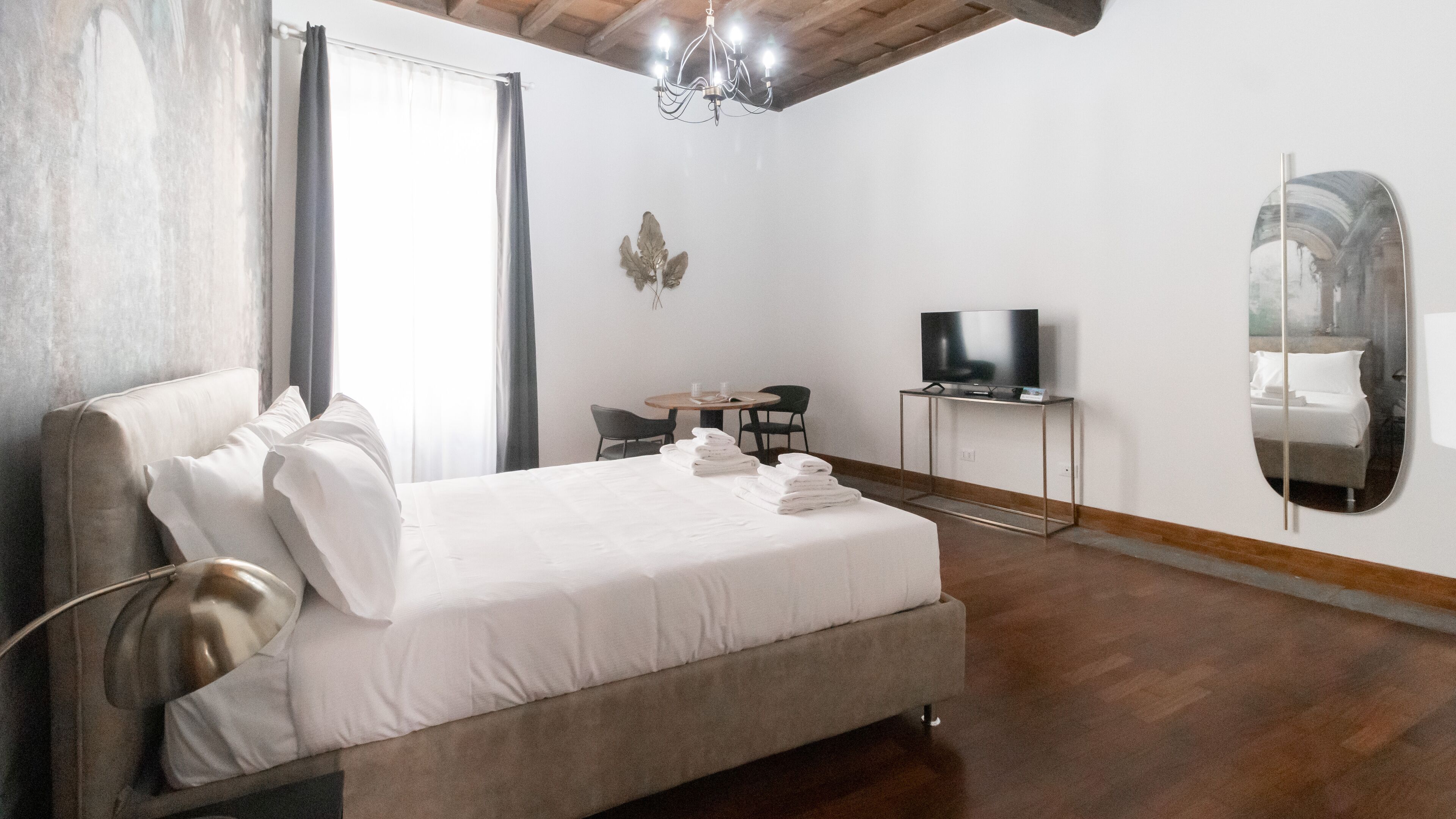 A bedroom with a bed, a TV on a stand, a mirror, and a table with chairs.