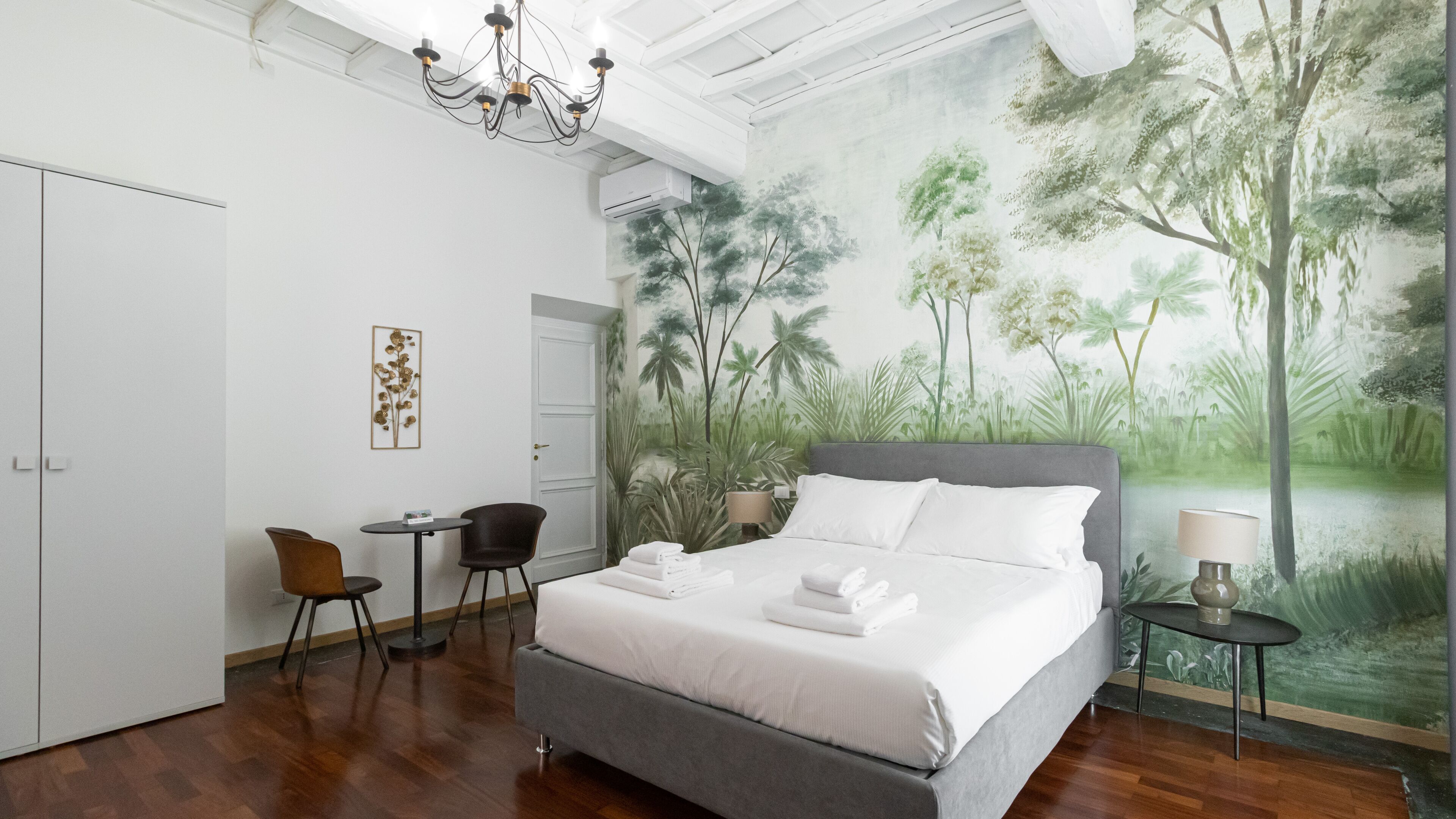 A bedroom with a large bed, a desk, two chairs, a chandelier, and a mural of a forest.
