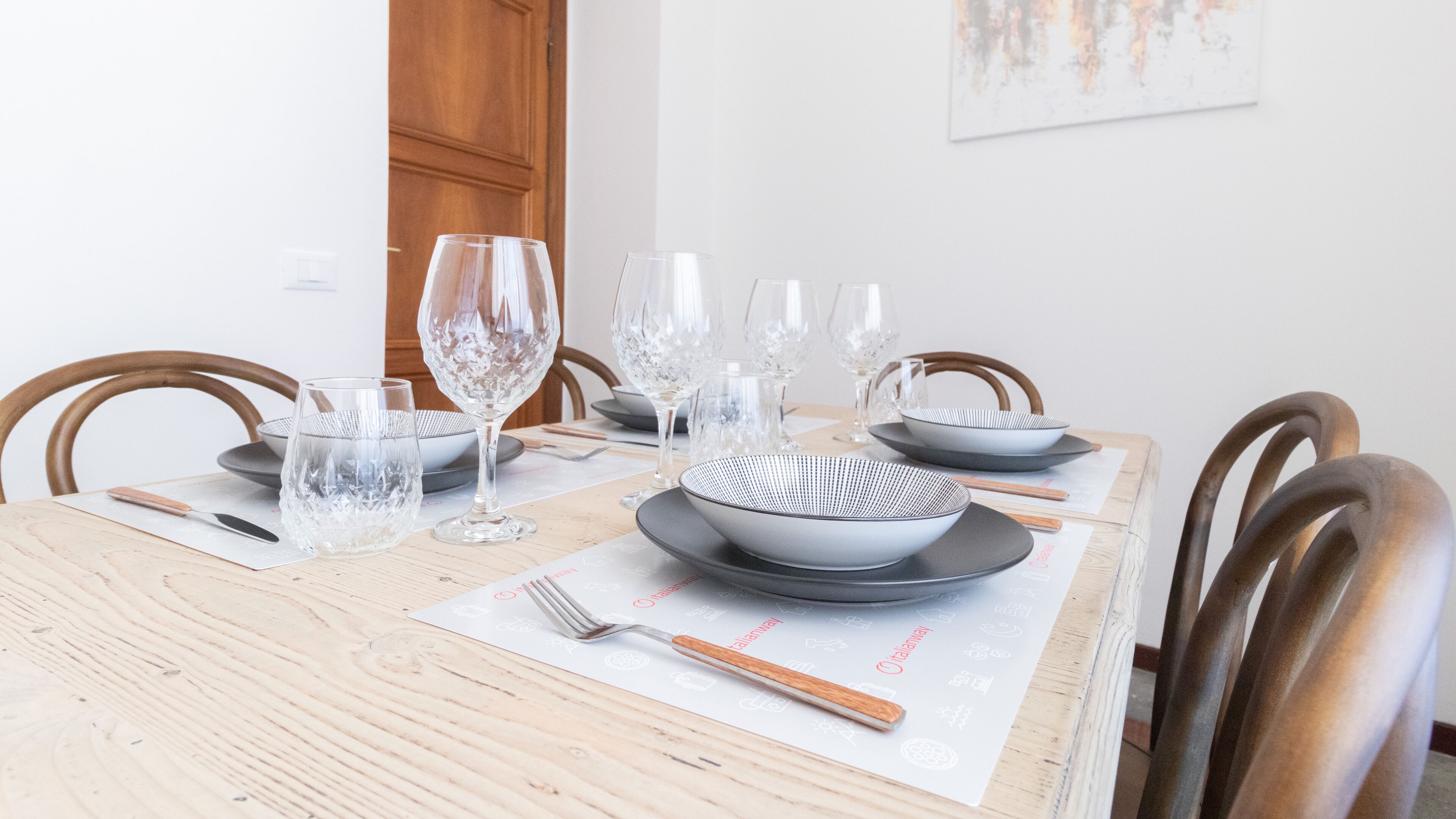 A dining table set with clear glassware, plates, and bowls, with a wooden chair and a door in the background.