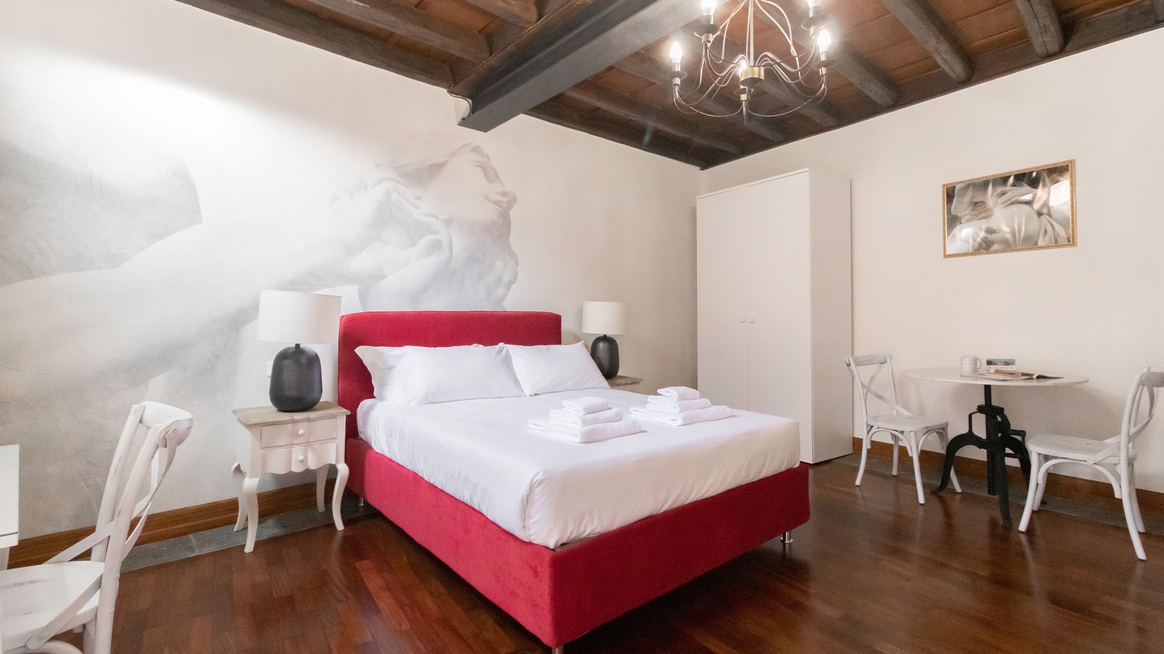 Une chambre d’hôtel avec un lit rouge, des murs blancs, un lustre, une petite table avec des chaises et un tableau accroché au mur.