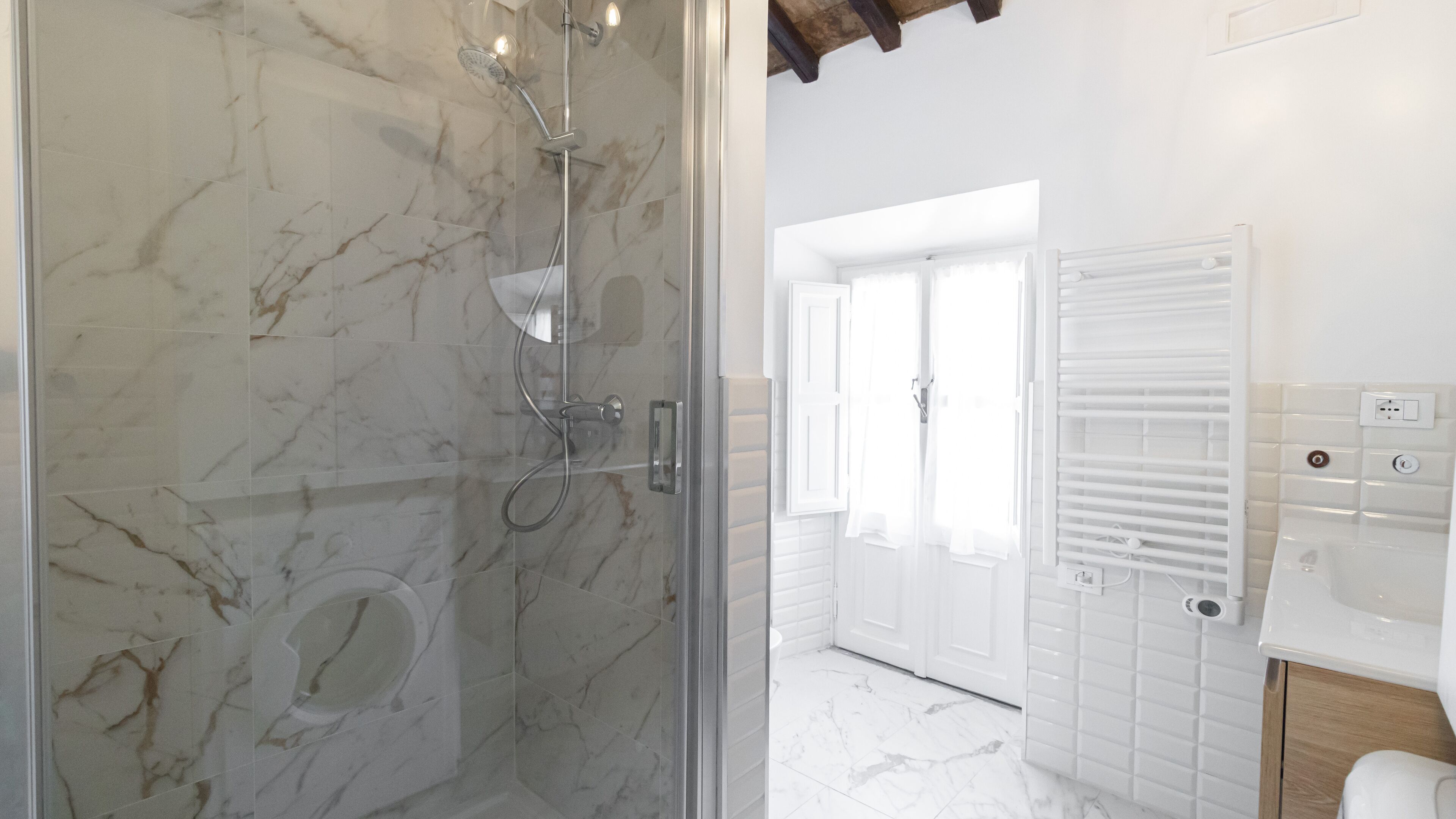 Une salle de bain dotée d’une douche à parois de verre, de murs recouverts de carreaux blancs, d’une baignoire blanche et d’un meuble-lavabo en bois avec un miroir.