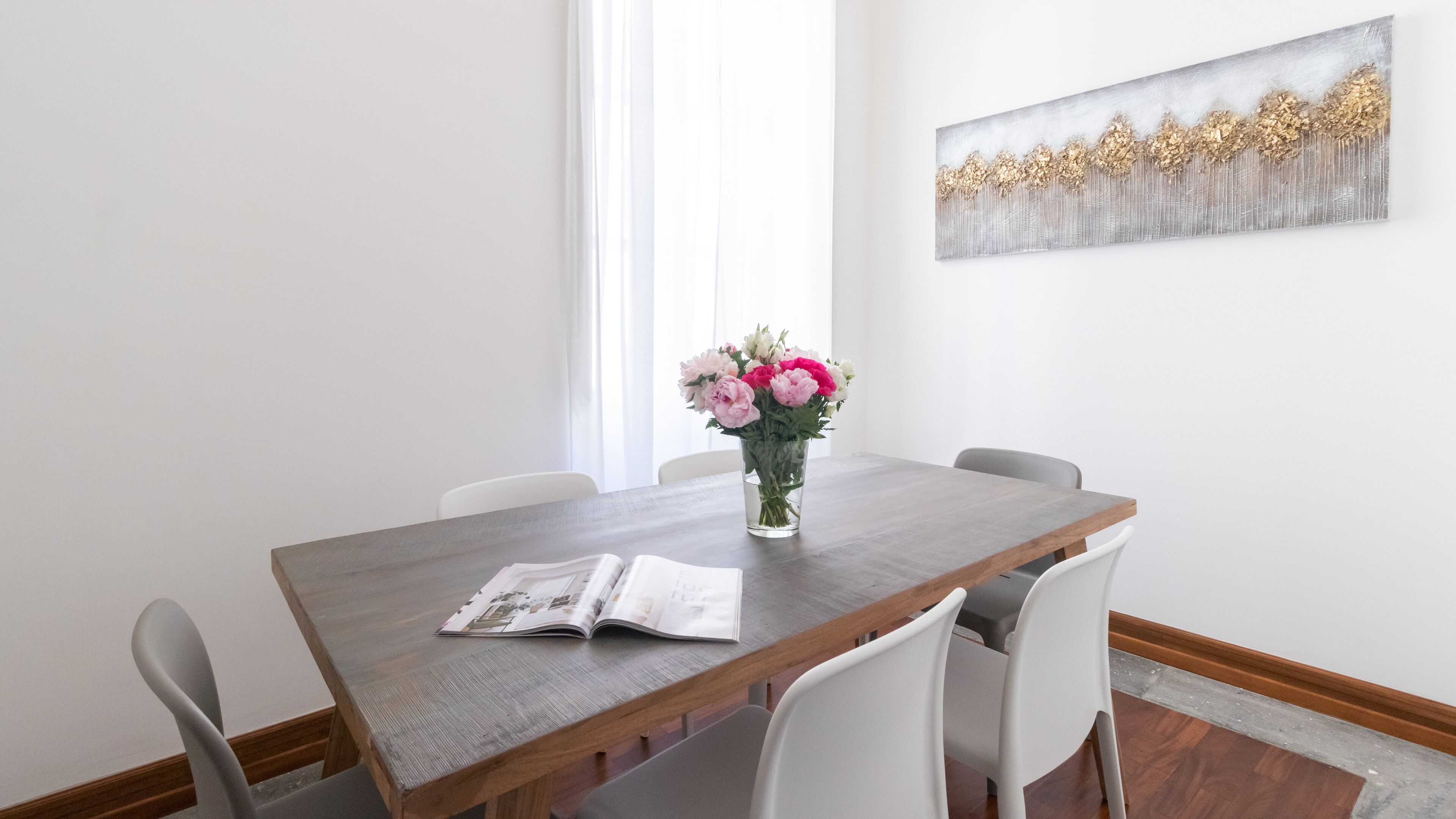 Un comedor con una mesa de madera, sillas blancas, un jarrón con flores y un cuadro en la pared.