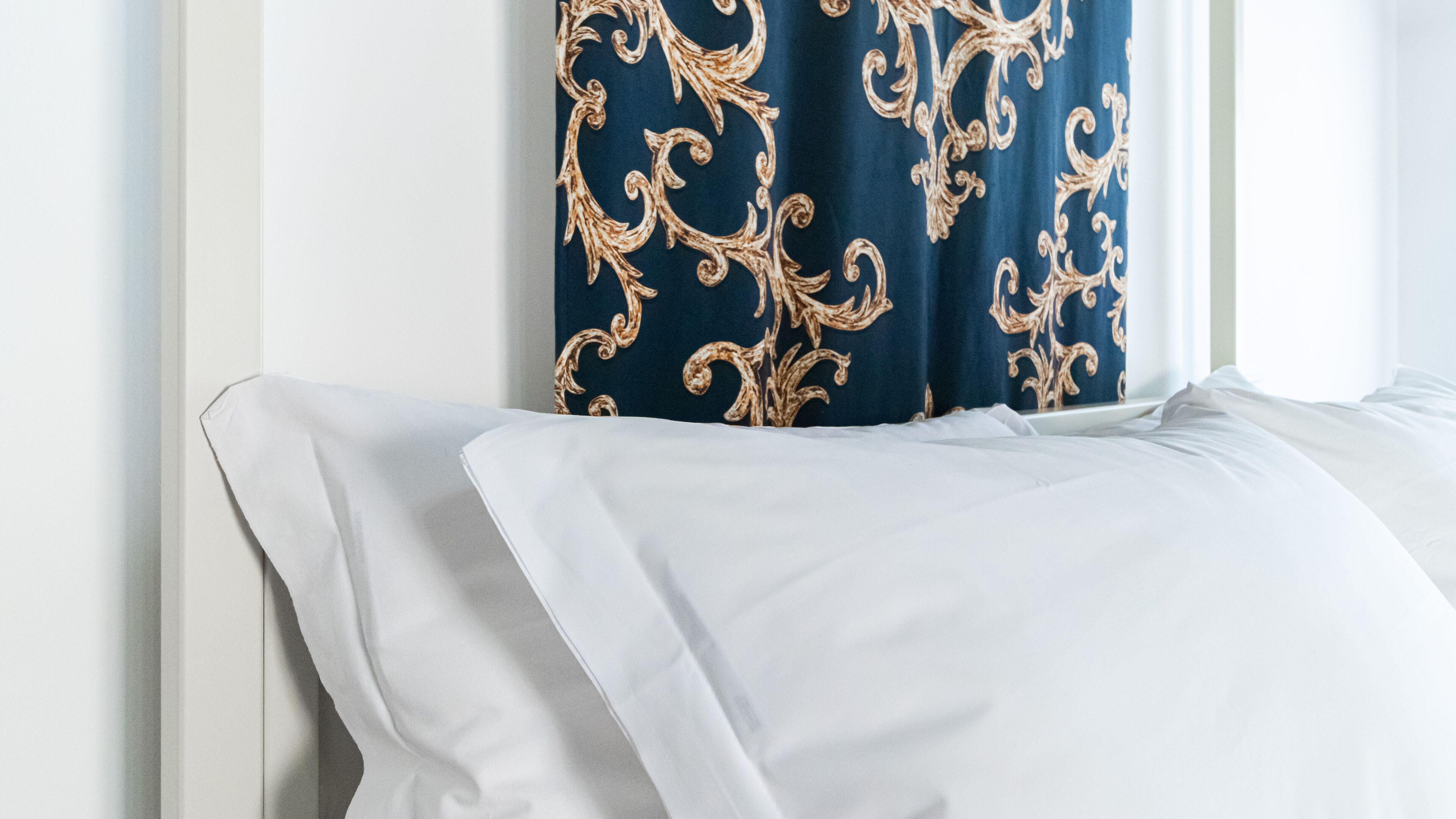 A bed with white linens and a blue curtain with gold patterns.