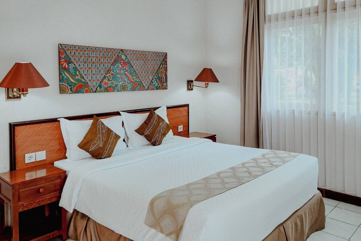A hotel room with a large bed, two bedside tables, a wall-mounted lamp, a decorative wall hanging, and a window with sheer curtains.