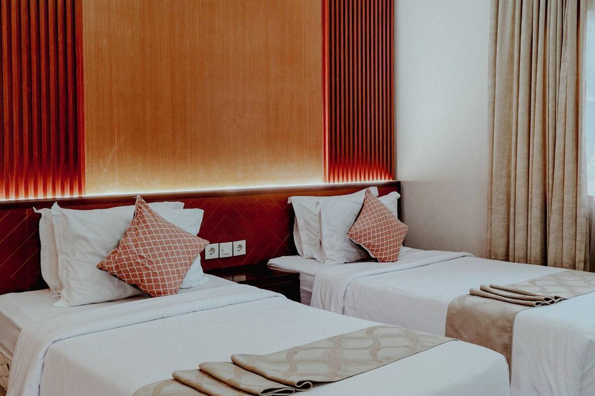 A hotel room with two beds, each with white linens and decorative pillows. The room features a wooden headboard, a large window with curtains, and a wall-mounted light switch.