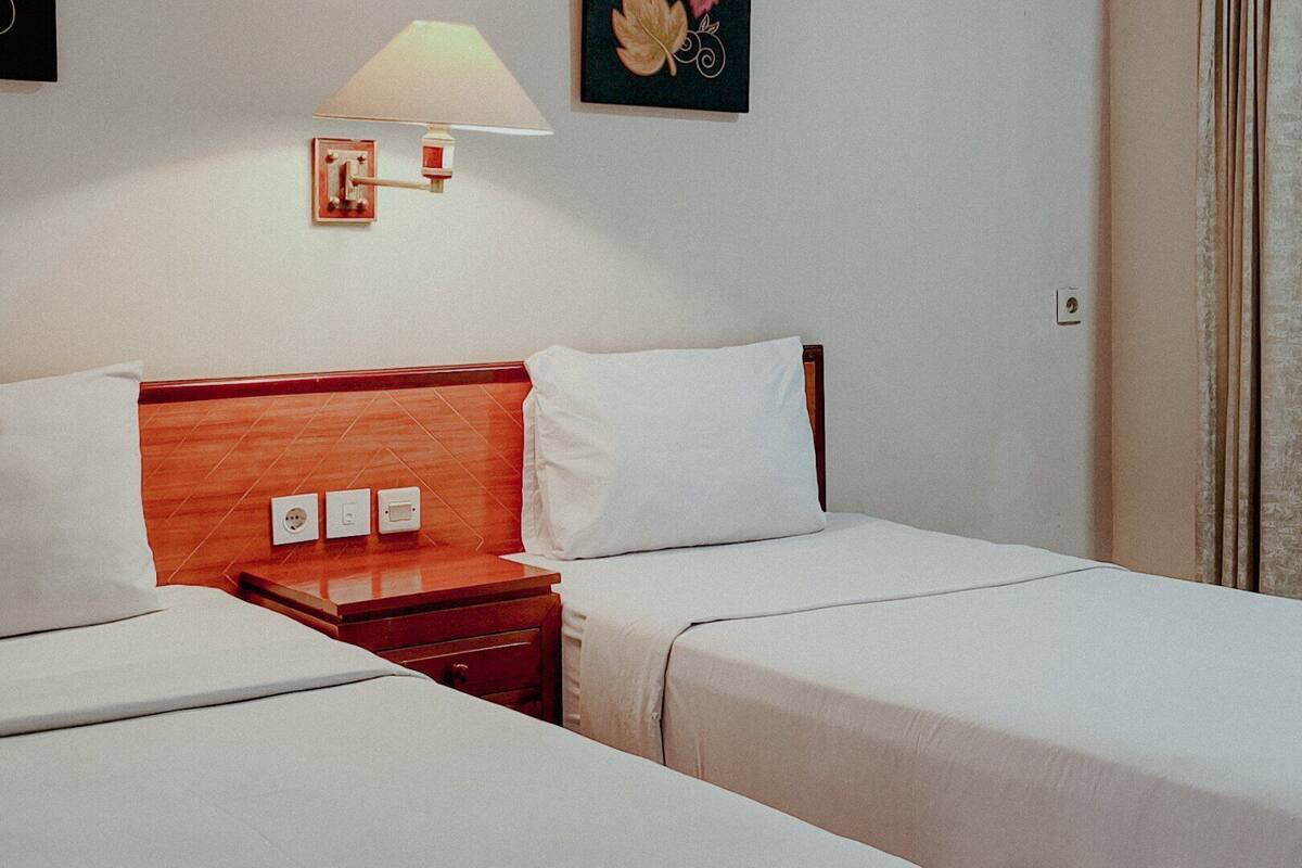A hotel room with two beds, a wooden headboard, a bedside table with a lamp, and a power outlet.