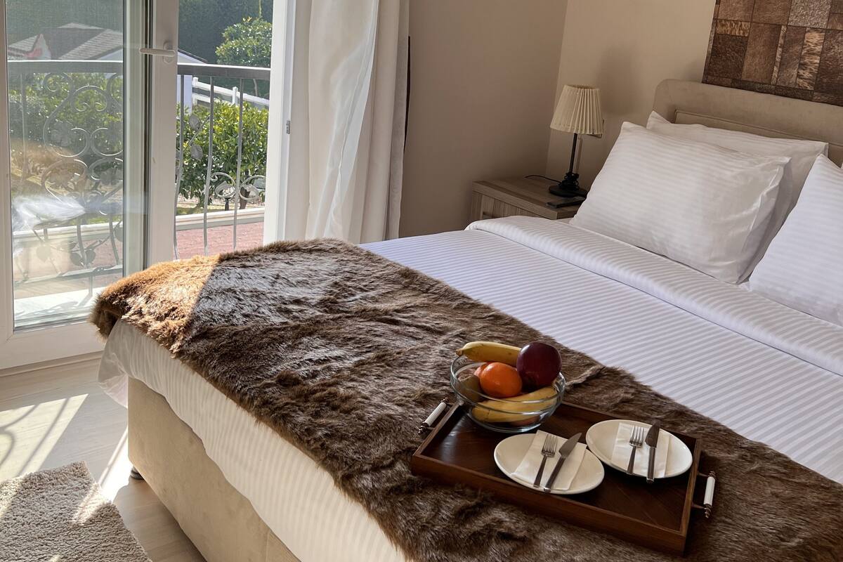 Une chambre avec un grand lit, une vue sur la verdure et les montagnes par la fenêtre, une tête de lit en bois et un plateau avec des fruits et des couverts posé sur le lit.