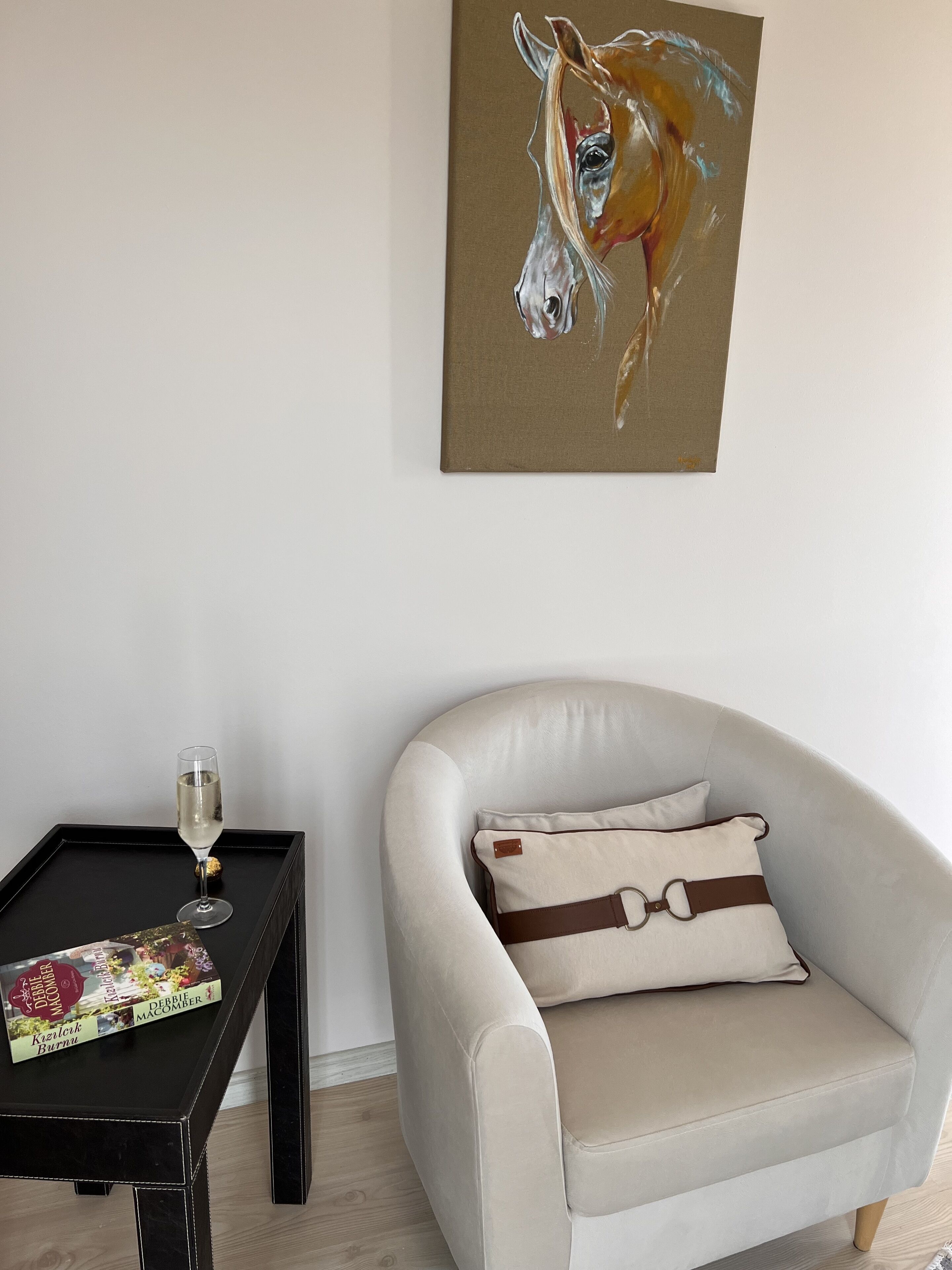 A room with a white armchair, a black side table with a glass of wine and a magazine, and a painting on the wall.