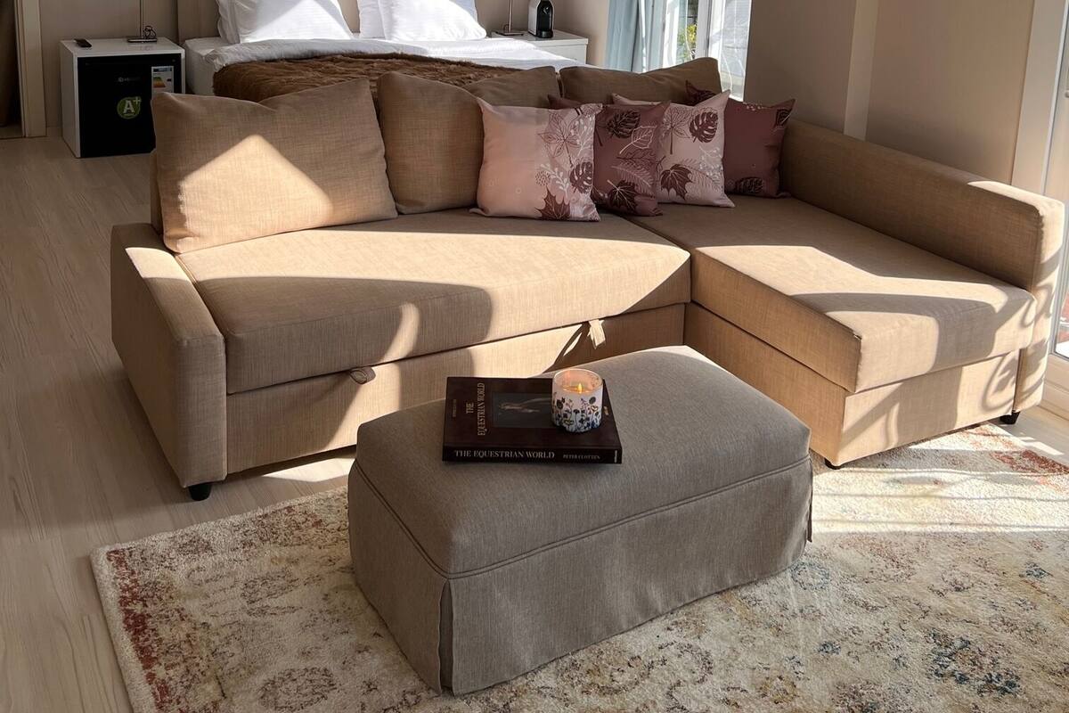 Une chambre d’hôtel moderne avec un grand canapé en forme de L, un petit pouf, une table de chevet avec une lampe et un lit doté d’une tête de lit.