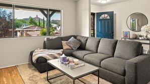Een woonkamer met een grijze hoekbank, een salontafel en uitzicht op een berg en huizen door een groot raam.