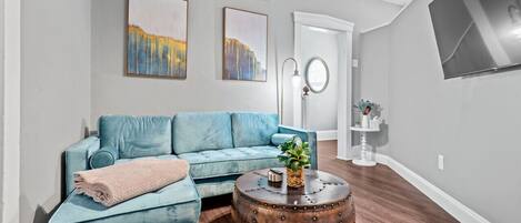 Una sala de estar con un sofá azul, una mesa redonda de centro con una planta y obras de arte en la pared.