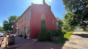 A red brick building with a green tree in front, a bench, and a paved street. - Historic Main St. Customs House Upstairs Apartment (Saint Charles)