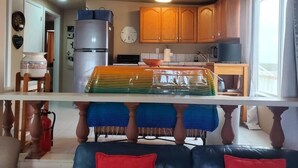 A compact kitchen with wooden cabinets, a refrigerator, a microwave, and a dining area with a table and chairs. - Hacienda De Sol y Luna- 2 bdrm Apartment (Playa Encanto)