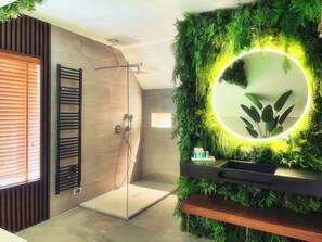Une salle de bain moderne avec une douche à l’italienne en verre, un meuble-lavabo en bois avec un miroir et un mur vert agrémenté d’un luminaire circulaire.