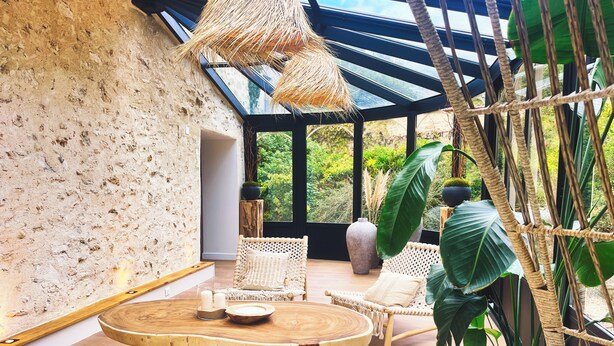 Une pièce avec une table ronde en bois, des chaises en osier, une grande plante en pot et une vue à travers un plafond en verre.