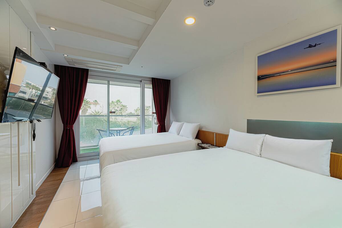 Ein Hotelzimmer mit zwei Betten, einem Fernseher, einem Sonnenuntergangsgemälde und einem Balkon mit Blick auf Palmen.