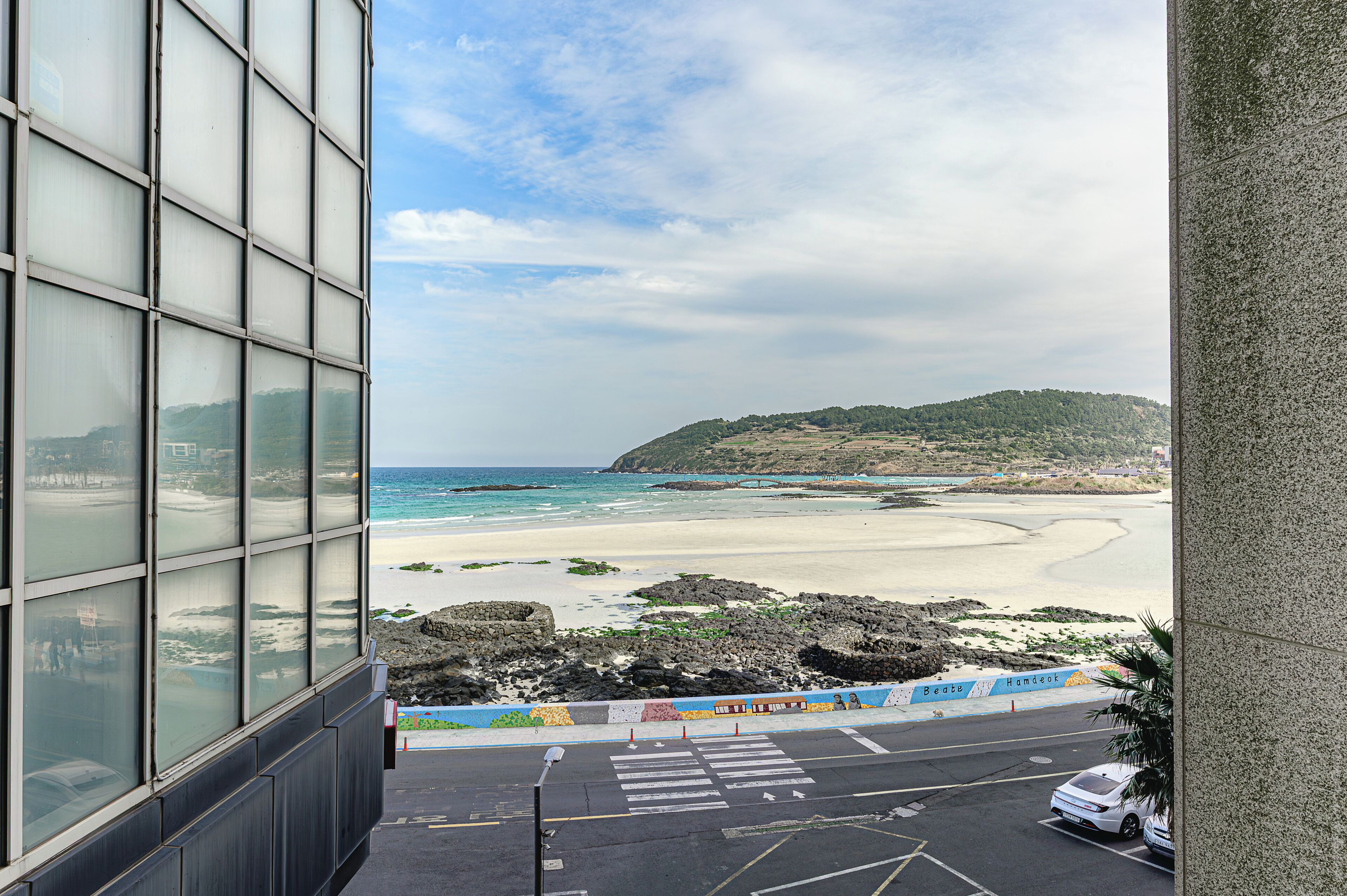 A view of a beach from a building with large windows, a parking area, and a clear sky.