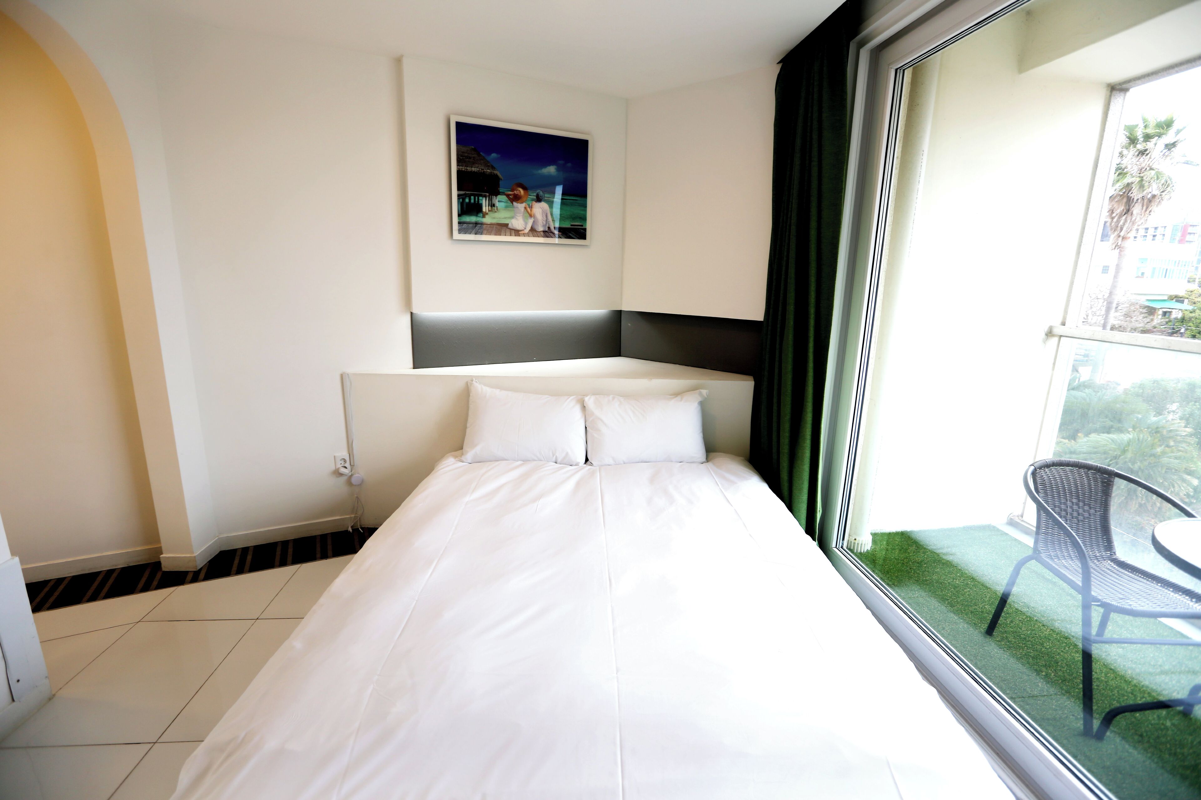 A hotel room with a bed, a painting on the wall, and a view of a balcony with a chair.