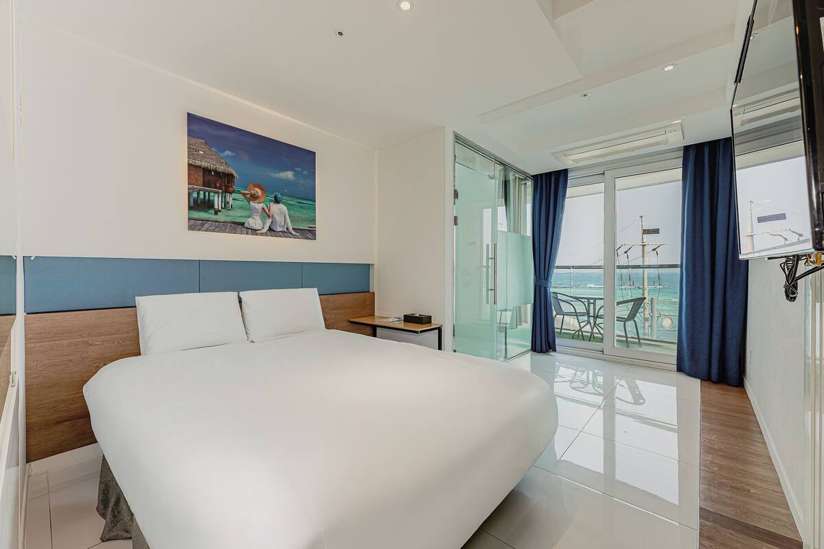Ein modernes Hotelzimmer mit einem großen Bett, einem kleinen Nachttisch, einem Gemälde an der Wand und einem Balkon mit Meerblick.