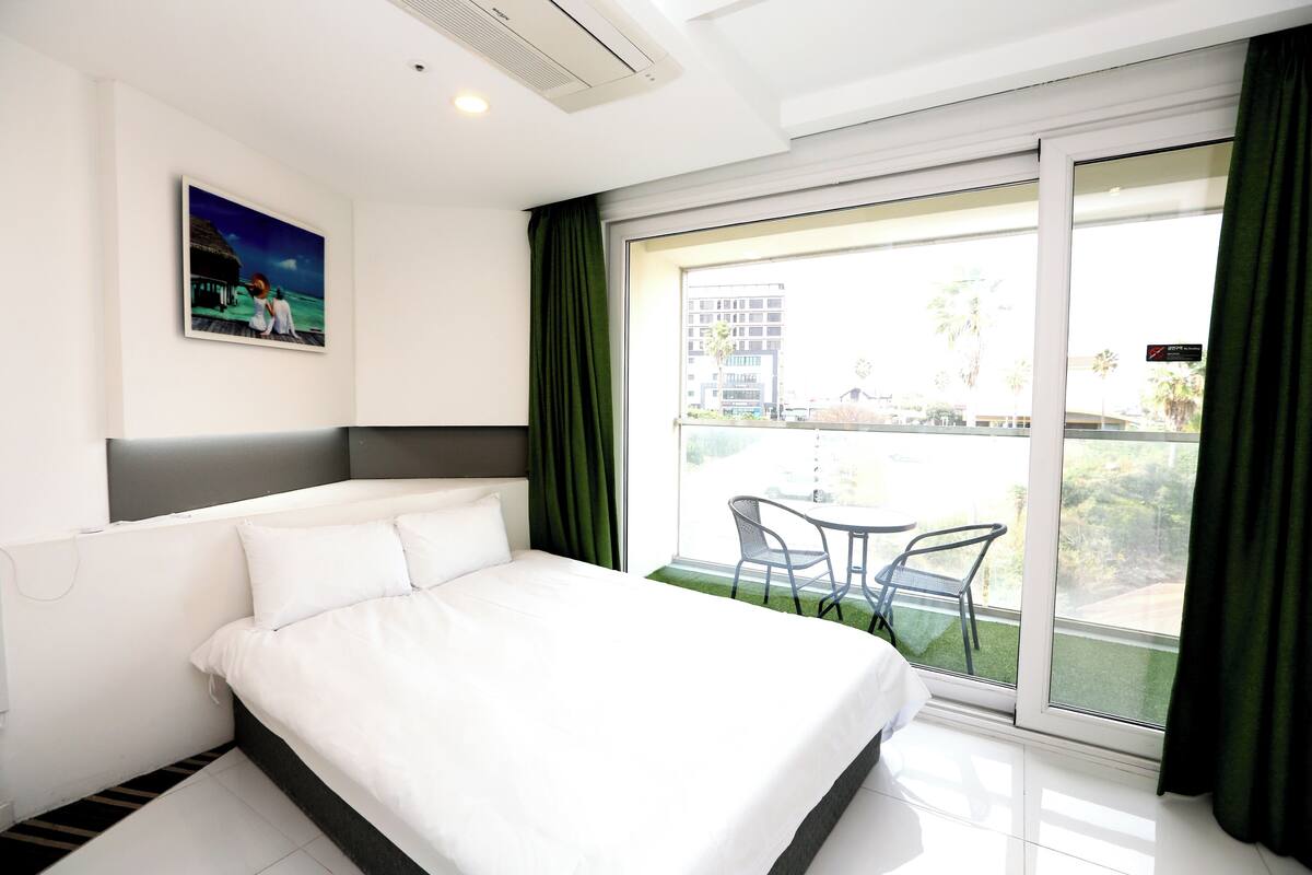 Ein Hotelzimmer mit Bett, einem Bild an der Wand, einem Balkon mit Tisch und Stühlen und Blick auf Gebäude und Palmen.