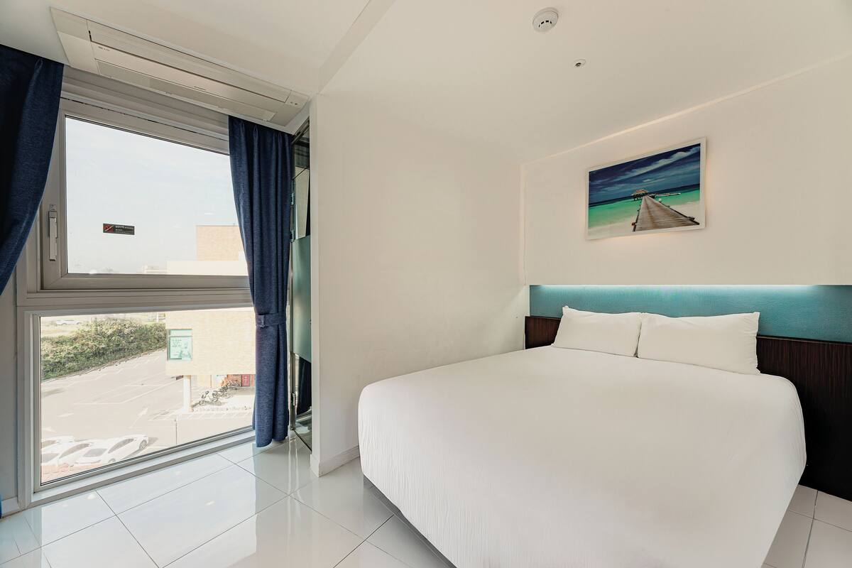 Ein Hotelzimmer mit einem großen Bett, einem Strandbild, einem Fenster mit Vorhängen und Blick nach draußen.