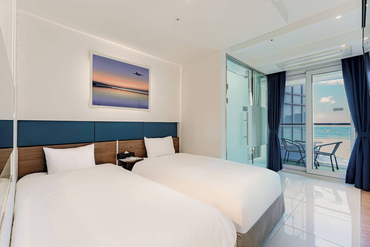 Ein Hotelzimmer mit zwei Betten, einem großen Fenster mit Blick auf den Strand und einem Gemälde eines Flugzeugs an der Wand.