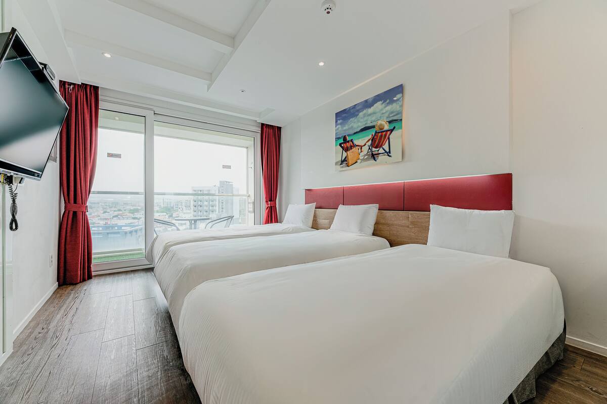 Ein Hotelzimmer mit zwei Betten, einem Fernseher, einem Gemälde und Blick auf die Stadt.