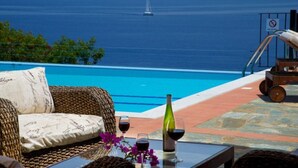 A poolside area with a wicker sofa, a table with a bottle and glasses, and a view of the sea with a sailboat. - Villa Alkionis in Kefalonia (Skala Kefalonia)