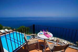 A balcony with a pool, wooden chairs, a table with books and a bottle, and a view of the sea. - Villa Alkionis in Kefalonia (Skala Kefalonia)