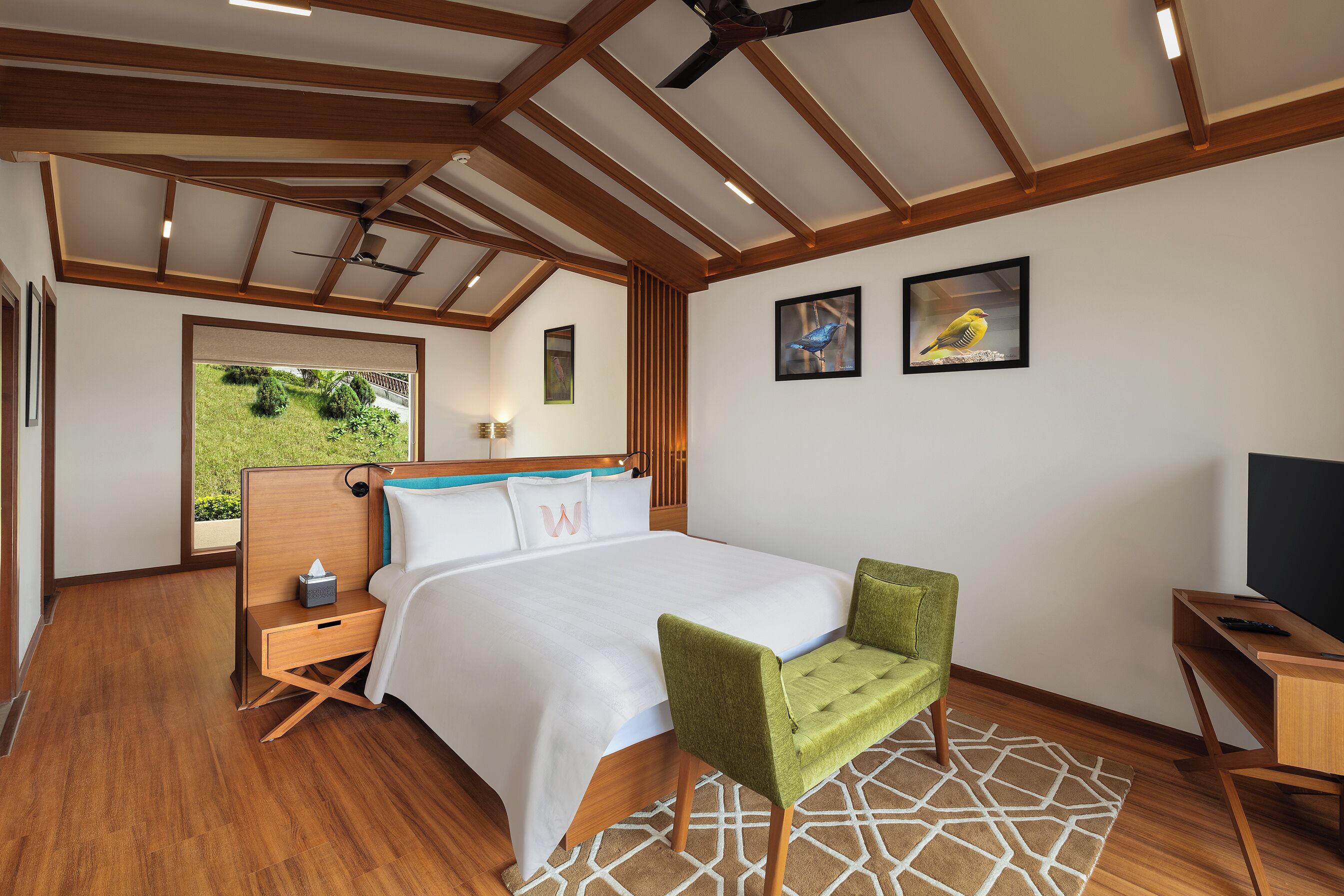 Une chambre d’hôtel moderne avec un plafond en bois, un lit recouvert de draps blancs, un fauteuil vert, un bureau et un téléviseur.
