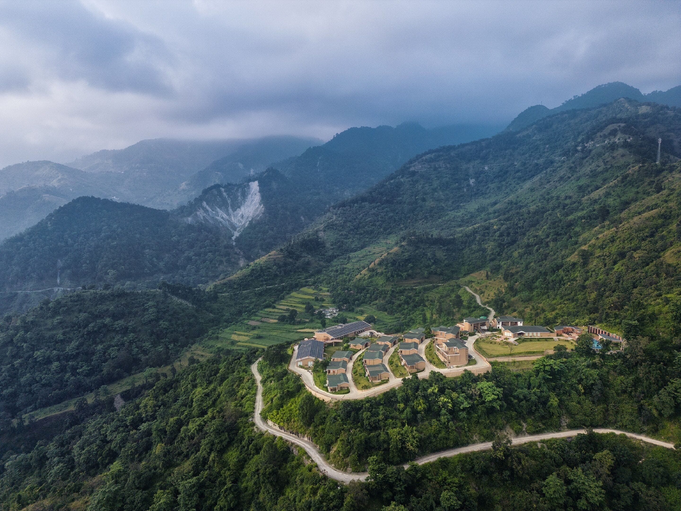A mountainous landscape with a residential area featuring houses with solar panels, surrounded by lush greenery and winding roads.