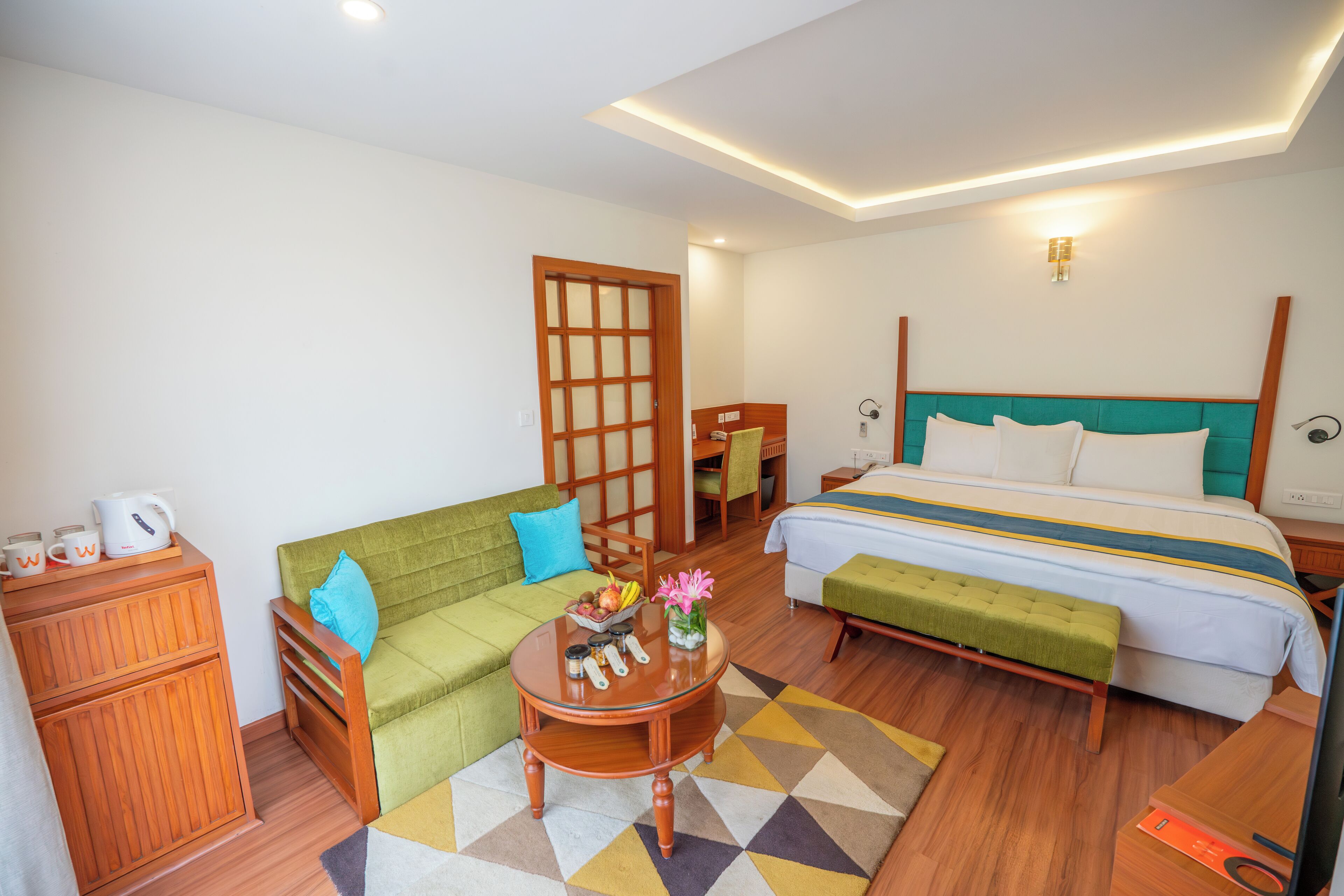 A hotel room with a bed, a sofa, a small table with a fruit bowl, a wooden cabinet, and a bench.