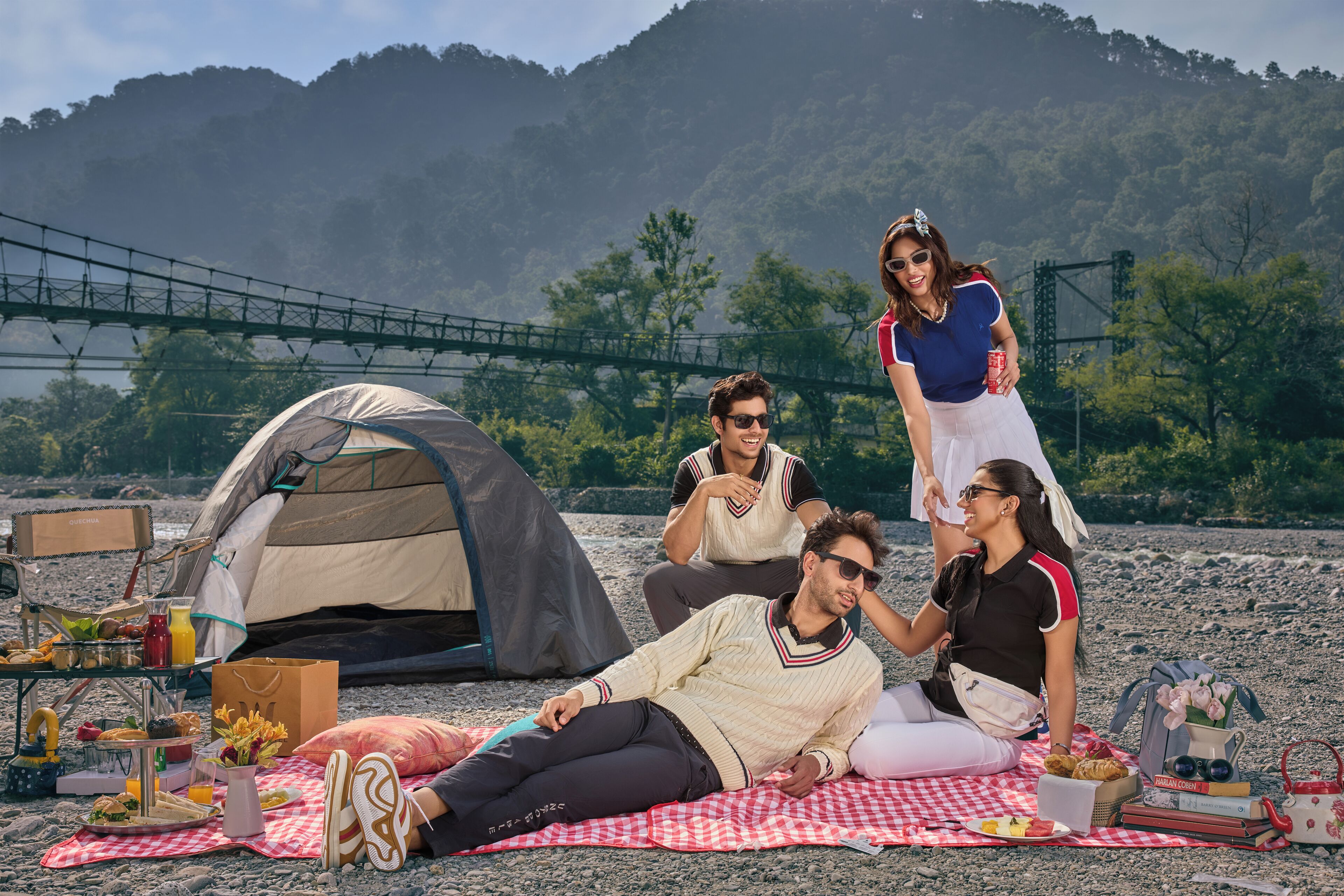 A group of people camping on a beach with a tent, picnic setup, and a bridge in the background.