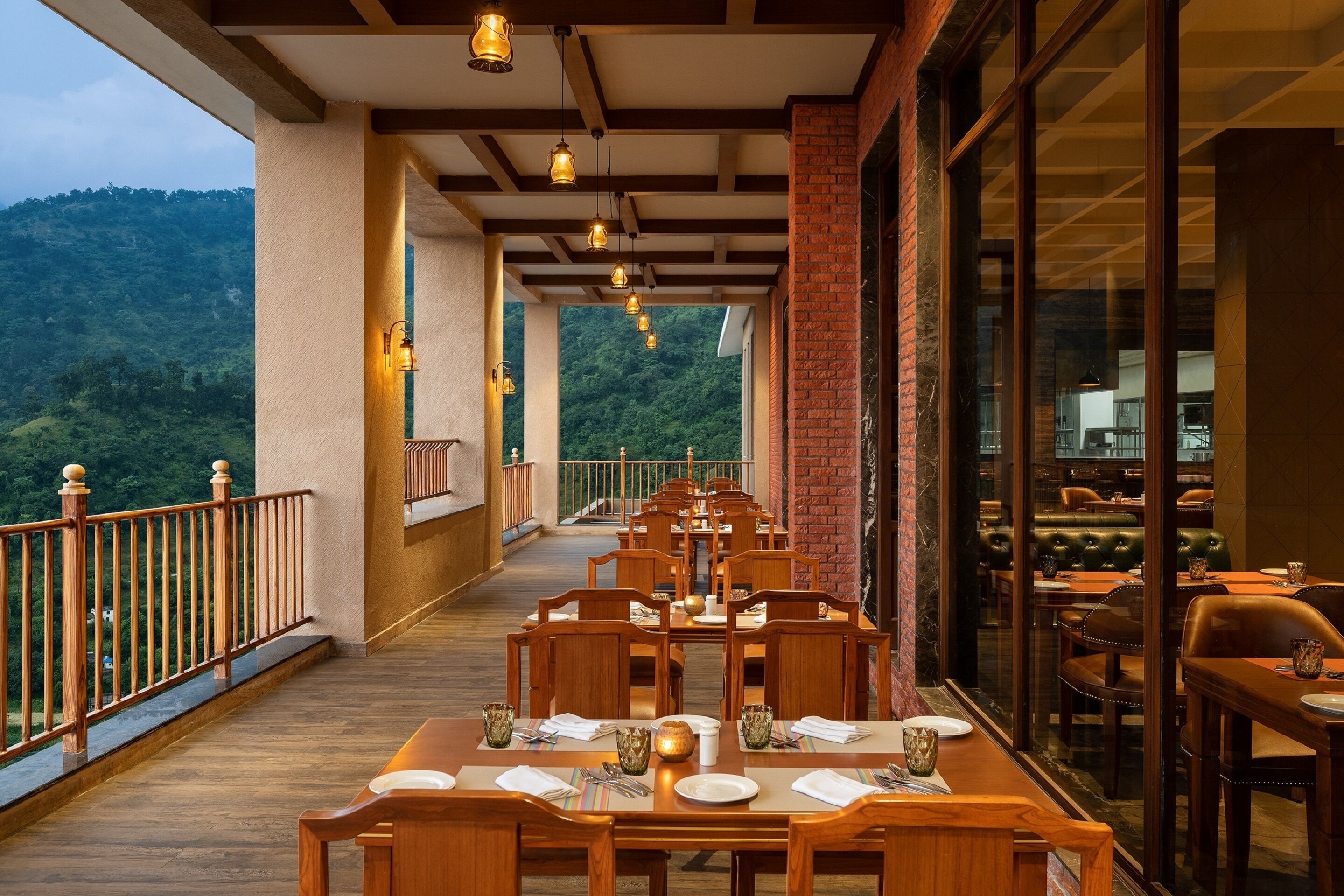 A restaurant with wooden tables and chairs, a balcony with a railing, and a view of a green hillside.