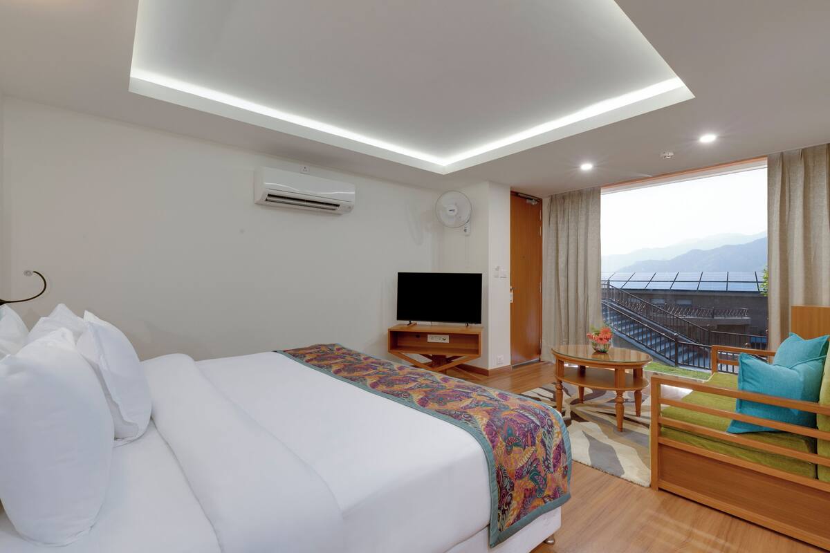 Habitación de hotel con una cama grande, televisión, balcón con vistas y una pequeña mesa con un jarrón de flores.