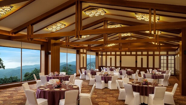 Une salle de banquet avec des tables rondes dressées pour le repas, de grandes fenêtres offrant une vue sur les montagnes, et un plafond haut orné de poutres en bois et de lustres.