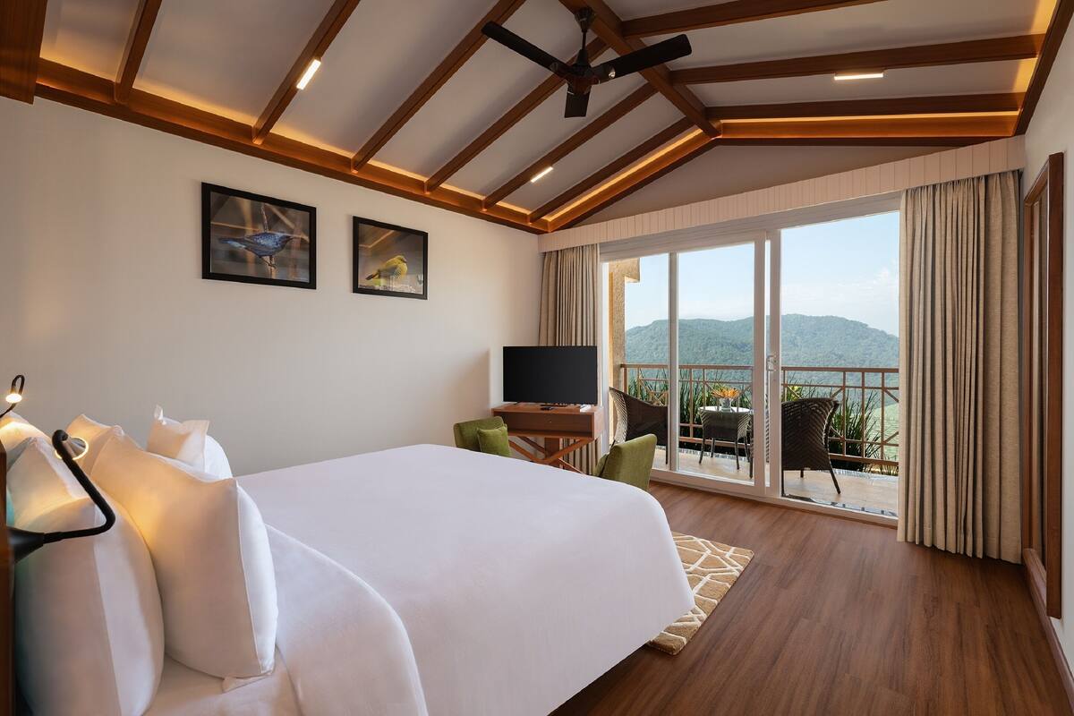 Habitación de hotel con cama, televisor, balcón con vista a las montañas y ventilador de techo.