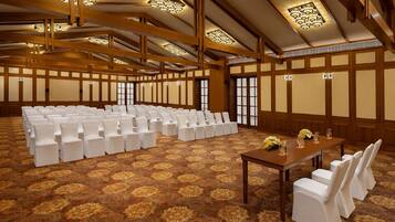 Un amplio salón de conferencias con filas de sillas blancas, una larga mesa con centros de mesa florales y paneles de madera en las paredes.