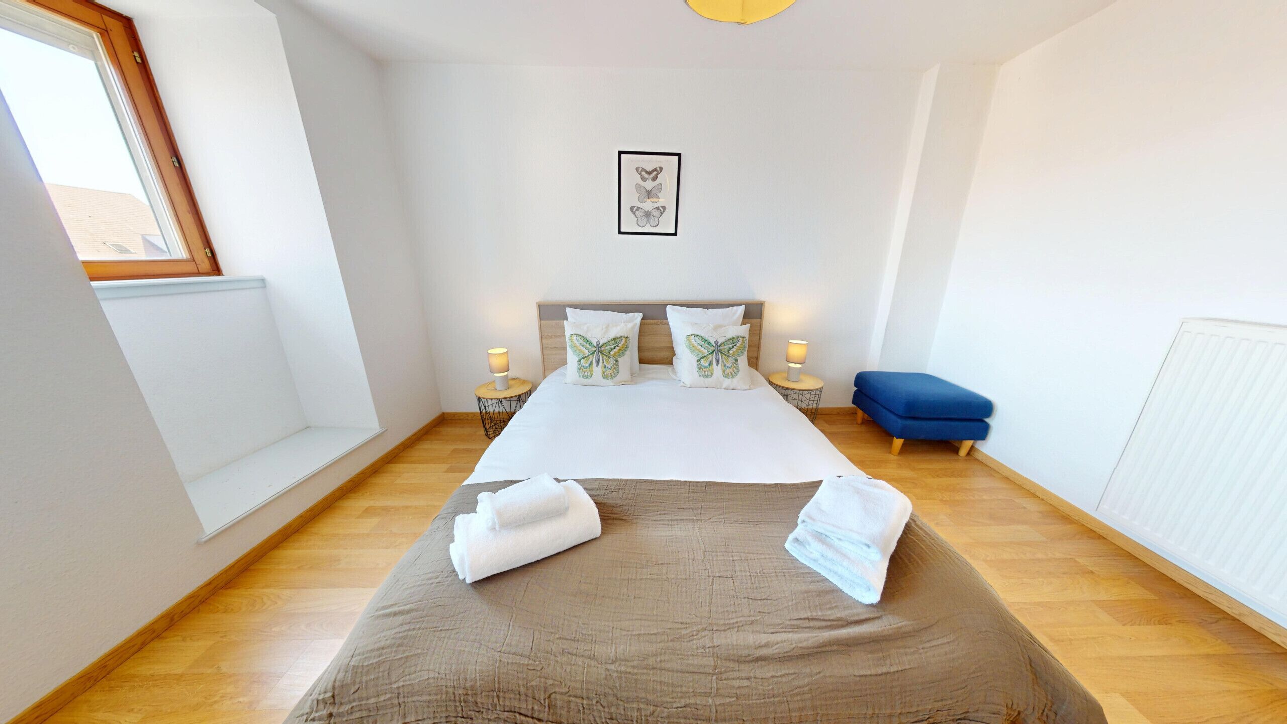 Una camera da letto con un letto grande, due comodini, una finestra, una panca blu e un quadro con farfalle incorniciate al muro.