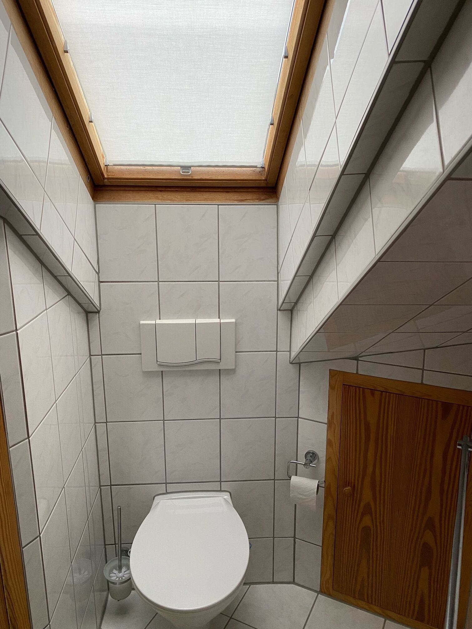 Ein modernes Badezimmer mit Oberlicht, weißen Fliesen, Toilette, Holztür und einer kleinen Mülltonne.