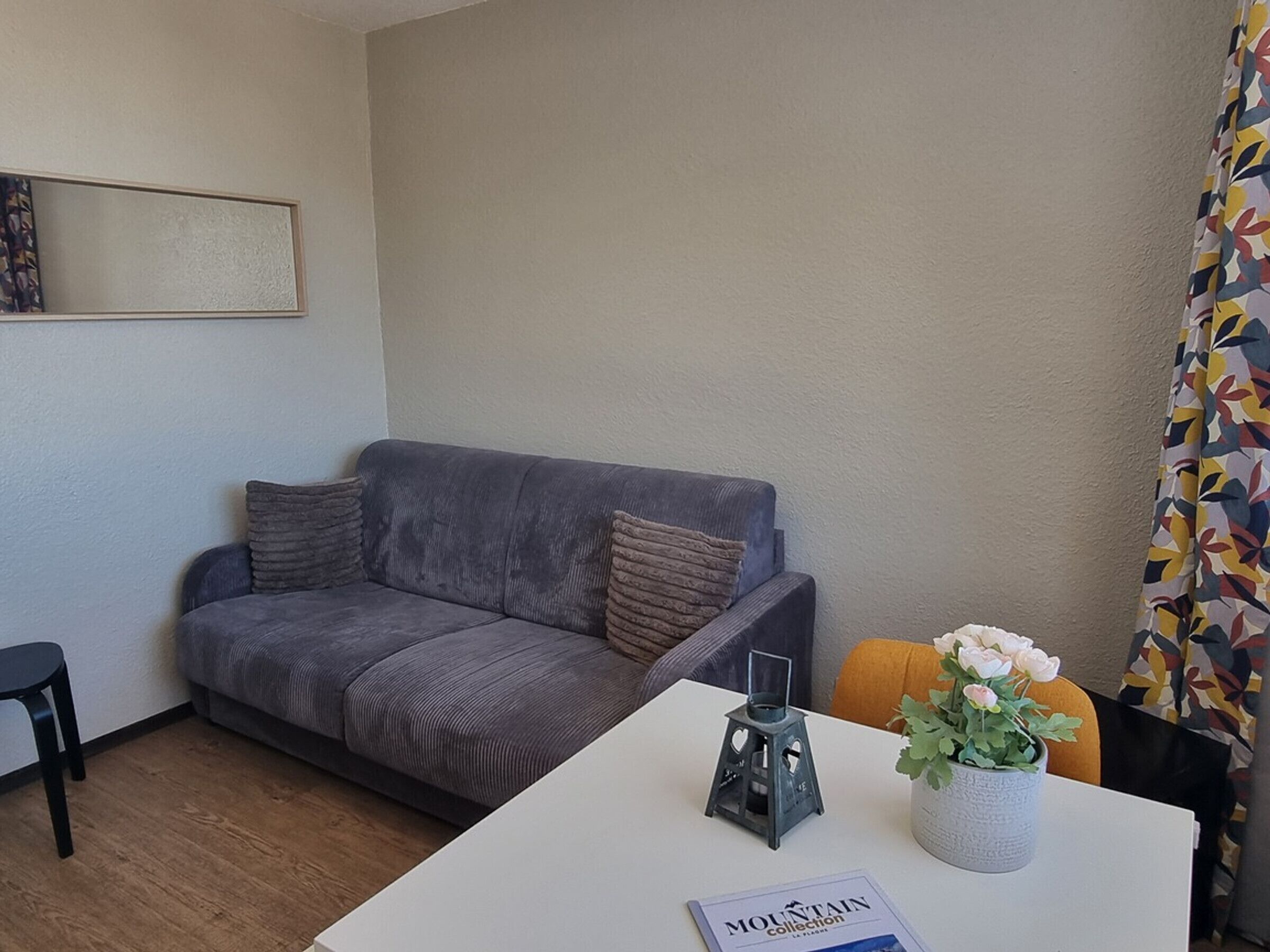 Un salon moderne avec un canapé gris, une petite table sur laquelle repose un livre, une plante décorative et un rideau coloré.