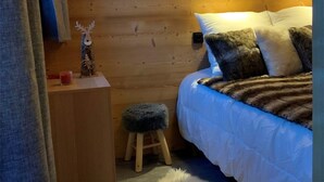 Un dormitorio acogedor con pared de madera, una cama con almohadas blancas y estampadas, un taburete pequeño y una figura decorativa de reno en un estante de madera.