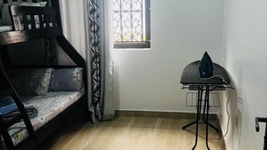 Een kamer met een stapelbed, een raam met decoratieve ijzeren spijlen, en een draagbaar strijkstation.