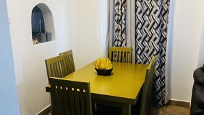 Een eetgedeelte met een gele tafel, vier houten stoelen en een zwarte schaal bananen.